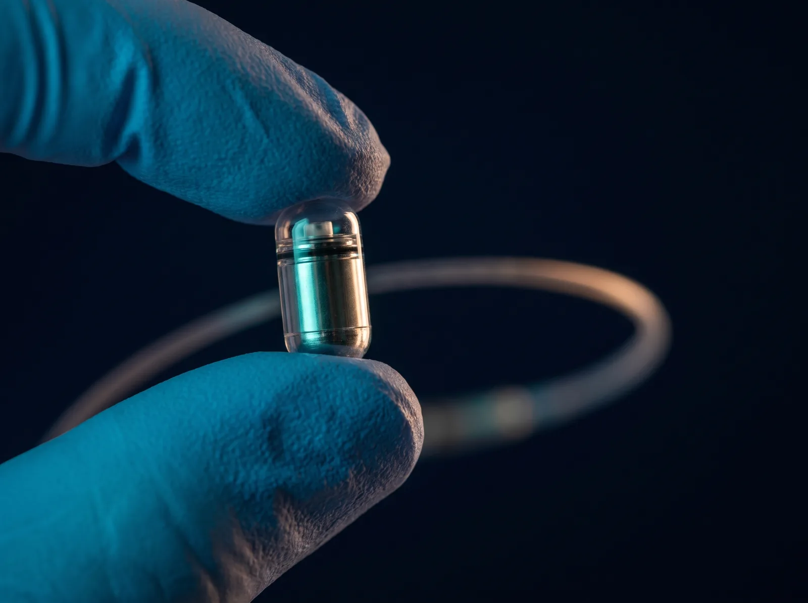 A gloved thumb and finger holding a small wireless pH capsule, with the delivery catheter coiled out of focus behind it.
