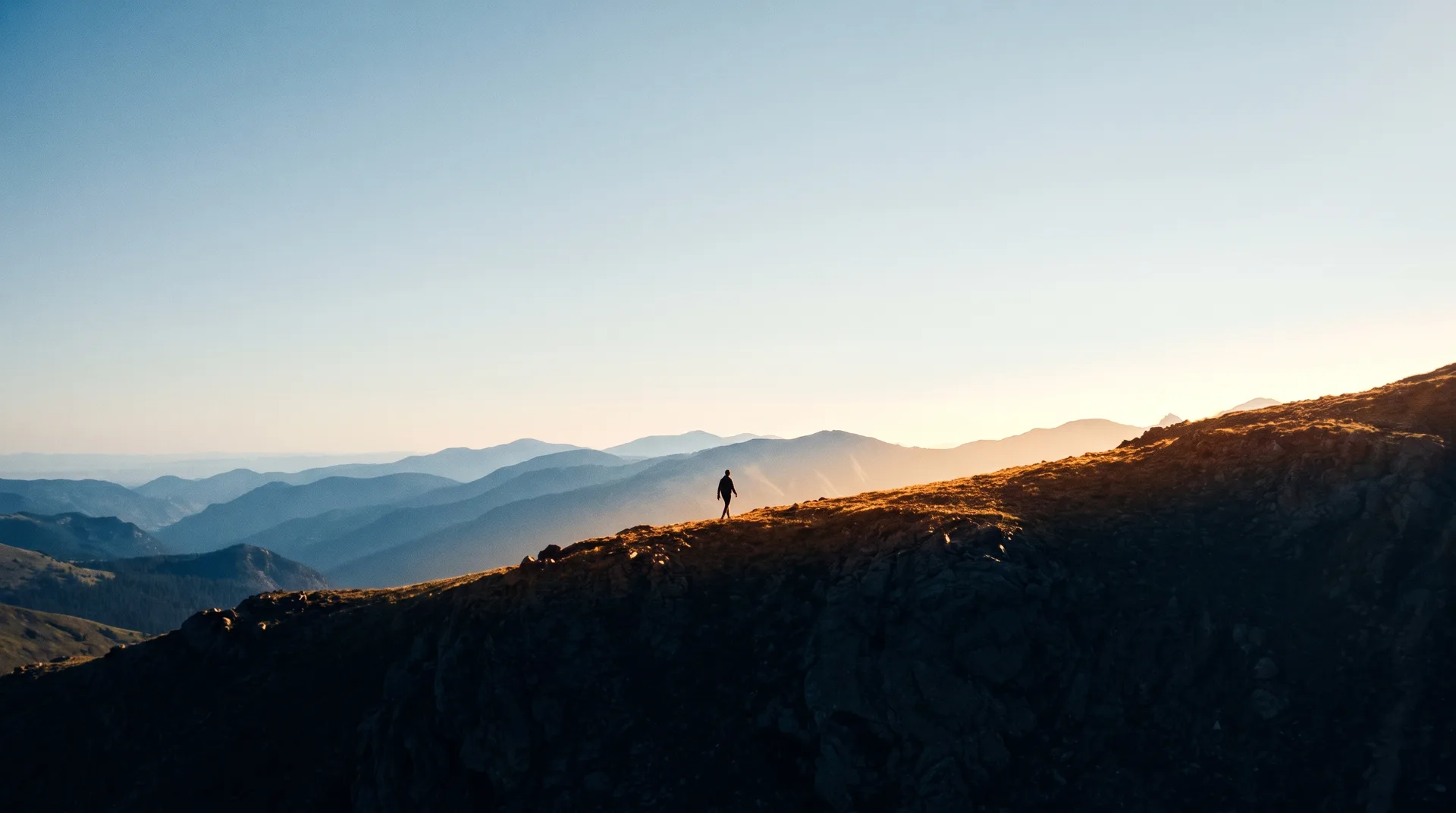 A single figure walking a high mountain ridge at sunrise above layered Colorado peaks.
